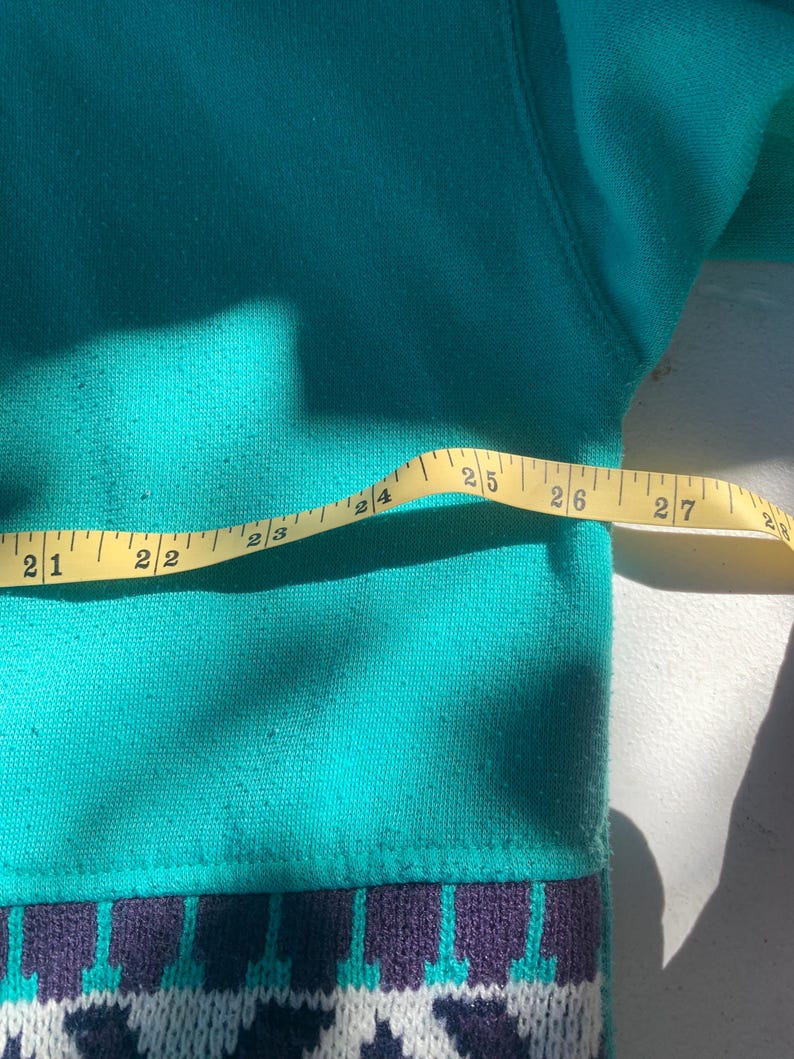Puede incluir: Un su&eacute;ter color teal con un borde inferior estampado. El borde presenta un dise&ntilde;o geom&eacute;trico en blanco, morado y teal. Una cinta m&eacute;trica amarilla se coloca sobre el su&eacute;ter, mostrando medidas de 53 a 69 cm.