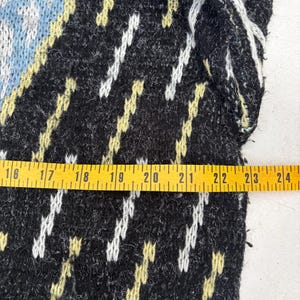 Puede incluir: Primer plano de una secci&oacute;n de un su&eacute;ter de punto negro con un patr&oacute;n geom&eacute;trico. El dise&ntilde;o incluye detalles en blanco, amarillo y azul claro. Una cinta m&eacute;trica amarilla se coloca sobre el su&eacute;ter.