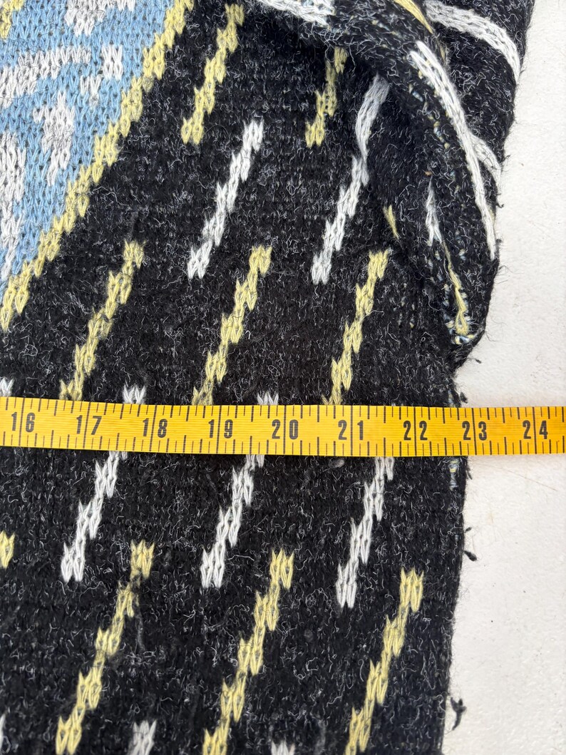 Puede incluir: Primer plano de una secci&oacute;n de un su&eacute;ter de punto negro con un patr&oacute;n geom&eacute;trico. El dise&ntilde;o incluye detalles en blanco, amarillo y azul claro. Una cinta m&eacute;trica amarilla se coloca sobre el su&eacute;ter.