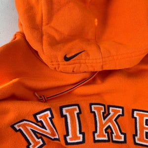 Peut inclure: Un sweat &agrave; capuche Nike orange vif avec cordon de serrage. Le mot "NIKE" est brod&eacute; sur le devant en blanc avec un contour bleu marine. Un petit logo Nike noir est sur la capuche.