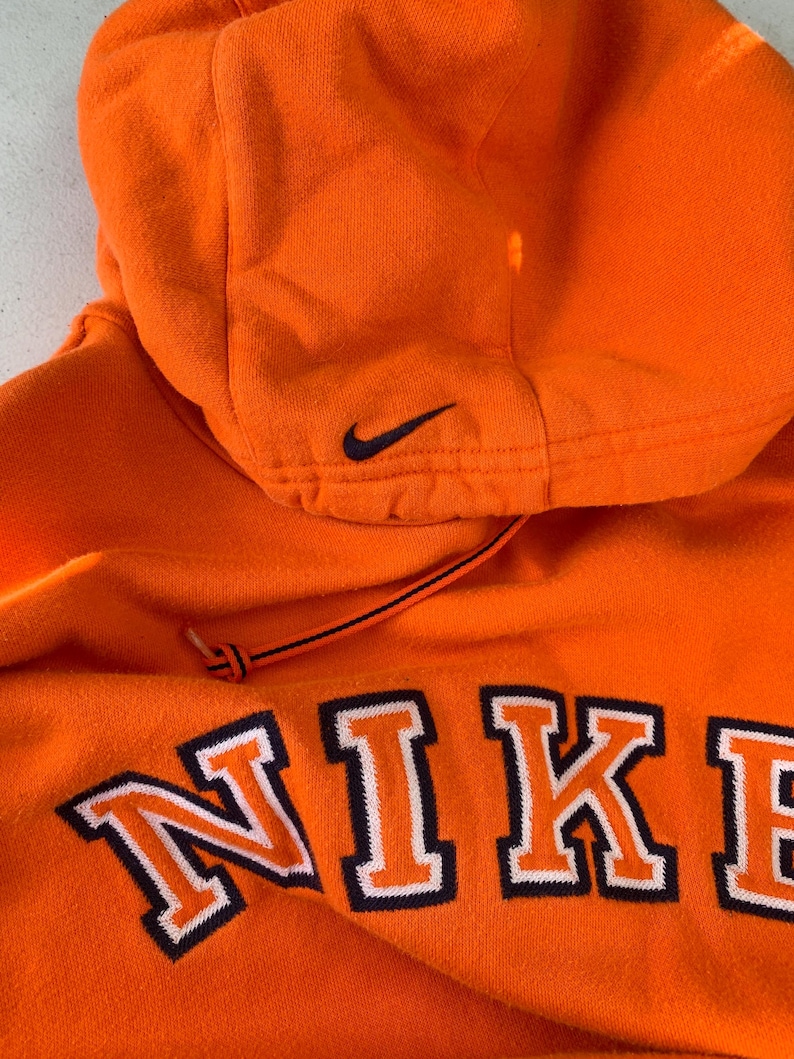 Peut inclure: Un sweat &agrave; capuche Nike orange vif avec cordon de serrage. Le mot "NIKE" est brod&eacute; sur le devant en blanc avec un contour bleu marine. Un petit logo Nike noir est sur la capuche.