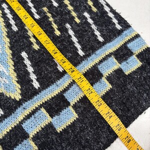 Puede incluir: Primer plano de una secci&oacute;n de una prenda de punto con un patr&oacute;n geom&eacute;trico. El dise&ntilde;o presenta colores negro, azul claro, blanco y amarillo. Una cinta m&eacute;trica amarilla se coloca sobre la tela, mostrando medidas en cent&iacute;metros.