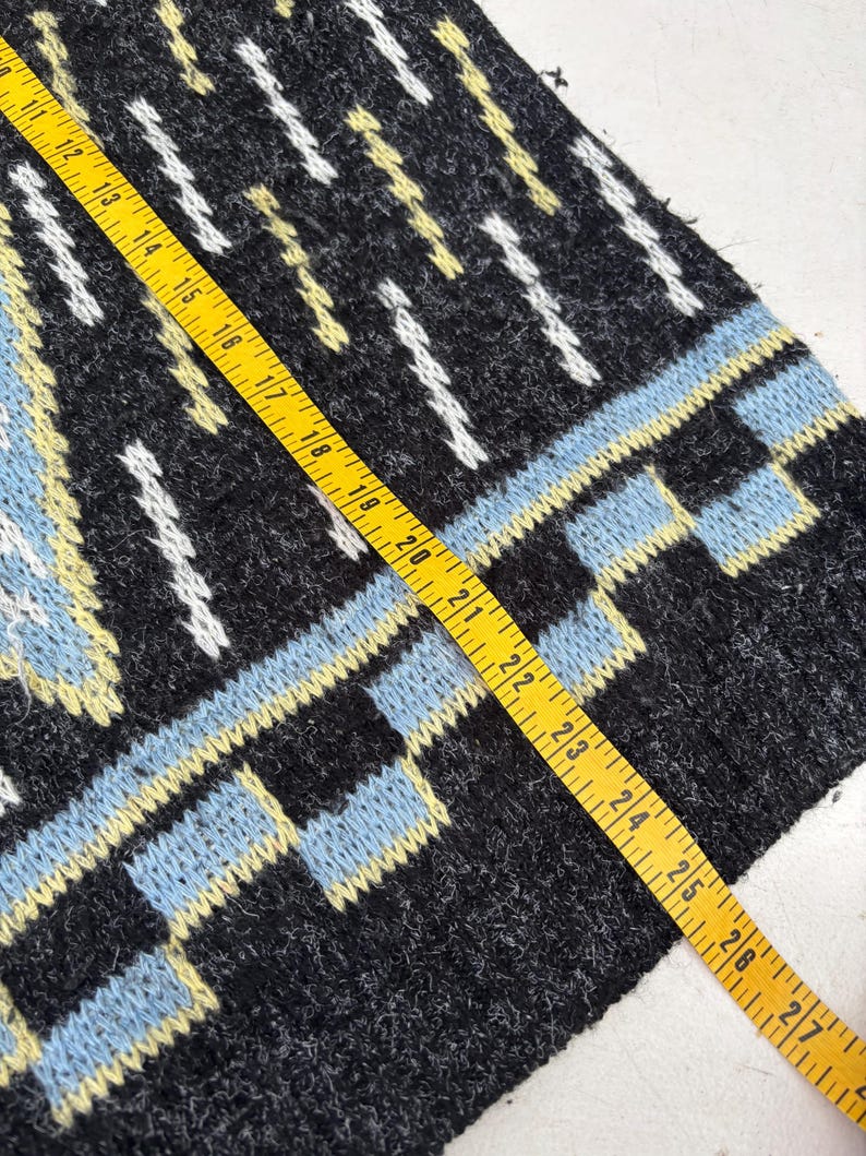 Puede incluir: Primer plano de una secci&oacute;n de una prenda de punto con un patr&oacute;n geom&eacute;trico. El dise&ntilde;o presenta colores negro, azul claro, blanco y amarillo. Una cinta m&eacute;trica amarilla se coloca sobre la tela, mostrando medidas en cent&iacute;metros.