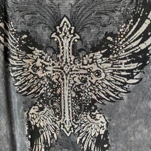Peut inclure: Un t-shirt &agrave; manches longues en maille gaufr&eacute;e grise avec un grand imprim&eacute; graphique noir et blanc repr&eacute;sentant une croix aux ailes d&eacute;ploy&eacute;es. Le motif est orn&eacute; de petits strass transparents et r&eacute;fl&eacute;chissants sur la croix et les ailes.