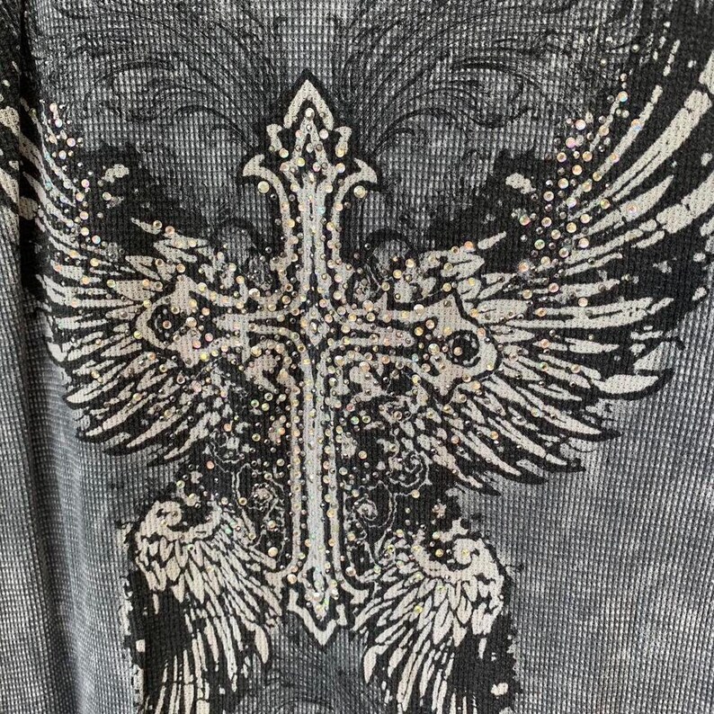 Peut inclure: Un t-shirt &agrave; manches longues en maille gaufr&eacute;e grise avec un grand imprim&eacute; graphique noir et blanc repr&eacute;sentant une croix aux ailes d&eacute;ploy&eacute;es. Le motif est orn&eacute; de petits strass transparents et r&eacute;fl&eacute;chissants sur la croix et les ailes.