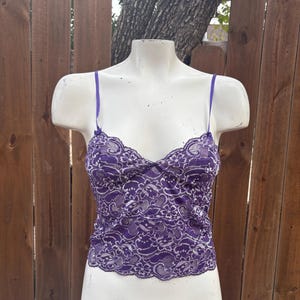 May include: A purple lace camisole with thin straps. The camisole is displayed on a white mannequin. The lace pattern is intricate, with a scalloped edge along the neckline and hem. The background is a wooden fence.
