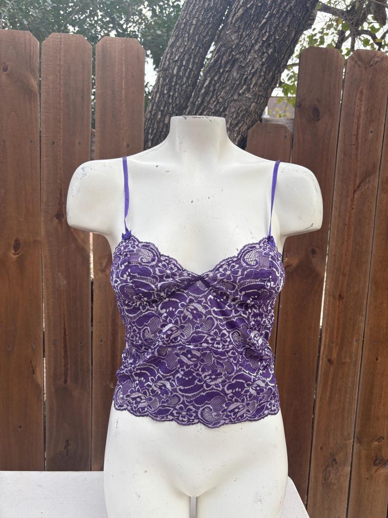 May include: A purple lace camisole with thin straps. The camisole is displayed on a white mannequin. The lace pattern is intricate, with a scalloped edge along the neckline and hem. The background is a wooden fence.