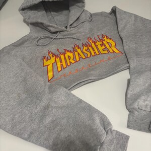 May include: A cropped gray hoodie with a drawstring hood and long sleeves. The front features the text "Thrasher Magazine" in yellow and orange flames. The sleeves have ribbed cuffs.