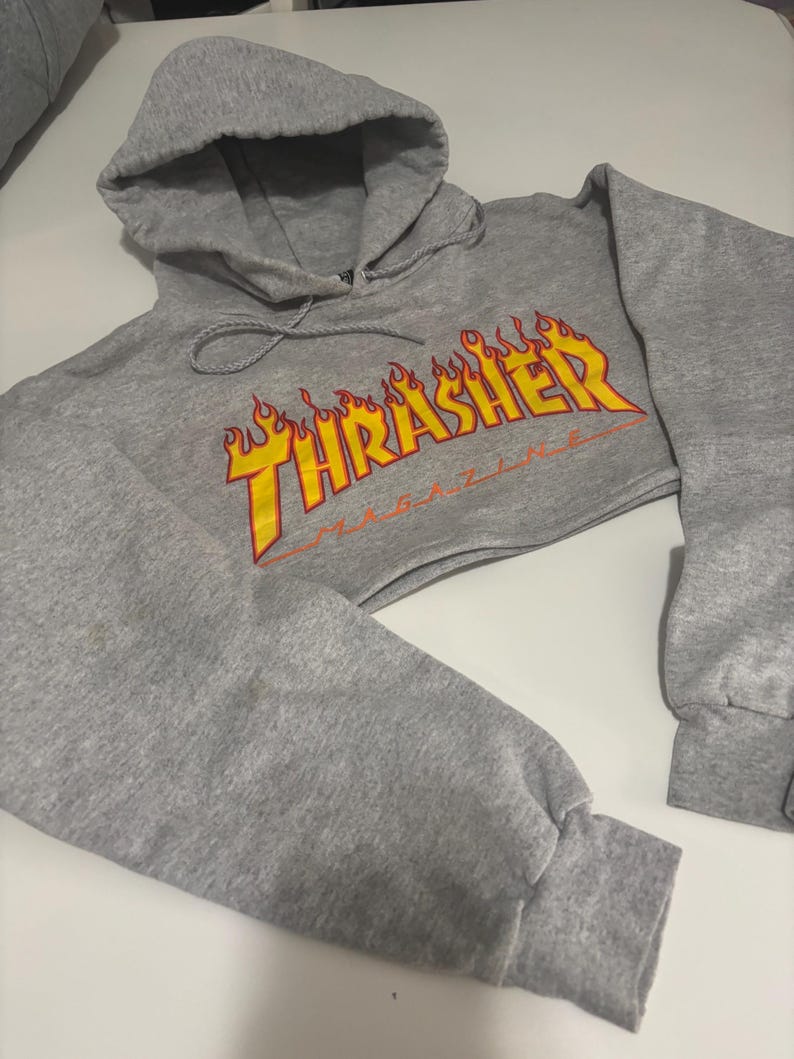 May include: A cropped gray hoodie with a drawstring hood and long sleeves. The front features the text "Thrasher Magazine" in yellow and orange flames. The sleeves have ribbed cuffs.