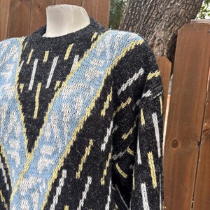 Puede incluir: Un su&eacute;ter gris con un patr&oacute;n geom&eacute;trico. El dise&ntilde;o incluye detalles en azul claro, amarillo y blanco. El su&eacute;ter tiene cuello redondo y mangas largas, perfecto para climas m&aacute;s frescos.
