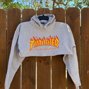 May include: A gray cropped hoodie with long sleeves. The front features the Thrasher Magazine logo in a flame design with red and yellow colors. The hoodie is hanging on a black hanger against a wooden fence.