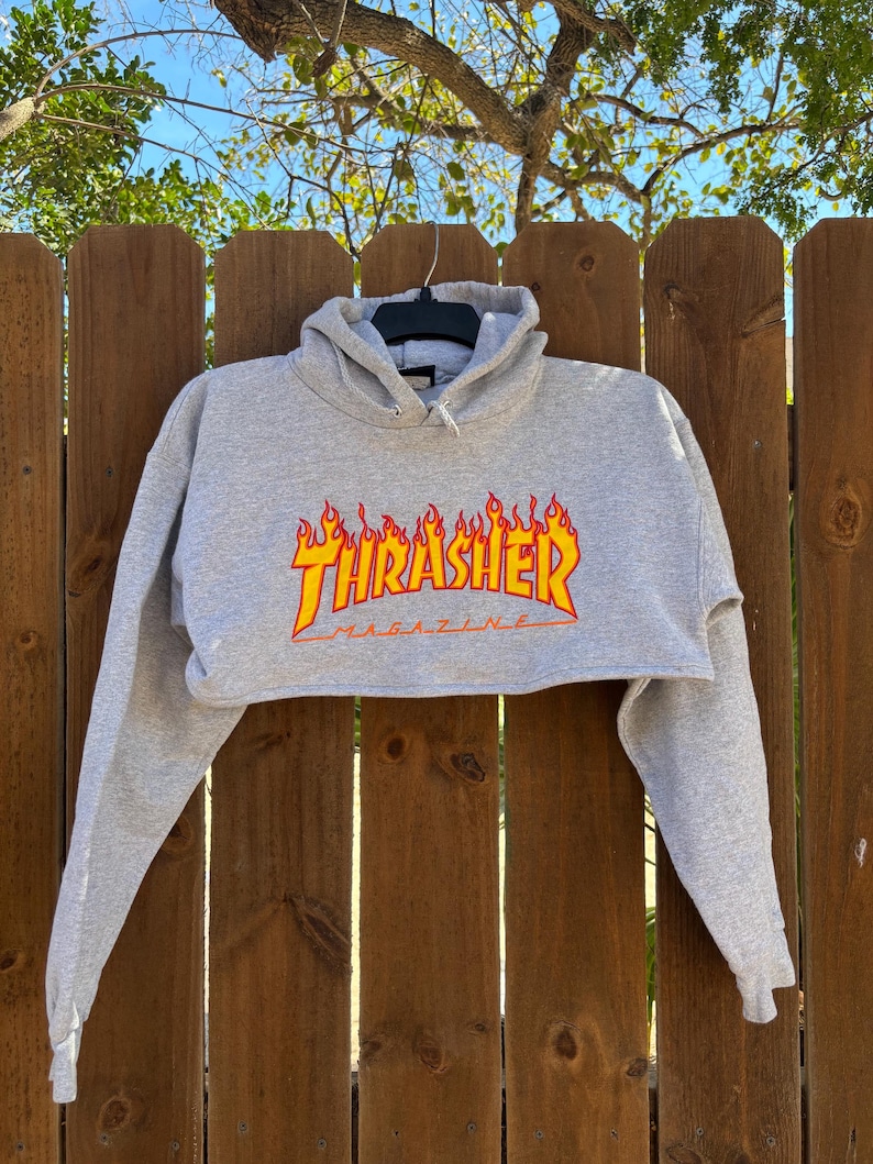May include: A gray cropped hoodie with long sleeves. The front features the Thrasher Magazine logo in a flame design with red and yellow colors. The hoodie is hanging on a black hanger against a wooden fence.