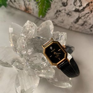 Pode incluir: Um relógio de pulso dourado com mostrador preto e pulseira de couro preta. O mostrador do relógio tem um diamante na posição das 12 horas e as palavras "BERBUS" e "DIA. SWISS 17 JEWELS" impressas nele.