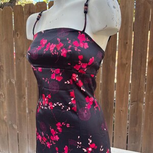 Op de afbeelding: Zwarte satijnen jurk met een bloemenpatroon in roze en rode tinten. De jurk heeft een vierkante halslijn, dunne bandjes met houten kralen en een getailleerd silhouet. Het bloemmotief bevat bloesems en Chinese karakters.