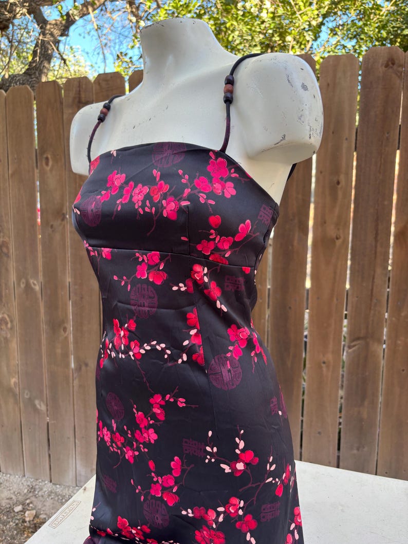 Op de afbeelding: Zwarte satijnen jurk met een bloemenpatroon in roze en rode tinten. De jurk heeft een vierkante halslijn, dunne bandjes met houten kralen en een getailleerd silhouet. Het bloemmotief bevat bloesems en Chinese karakters.