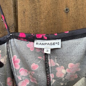 Op de afbeelding: Close-up van een zwarte jurk met een roze bloemenpatroon. De jurk heeft dunne bandjes en een V-hals. Het label leest "RAMPAGE USA" en maat "3". De stof heeft een subtiele textuur.