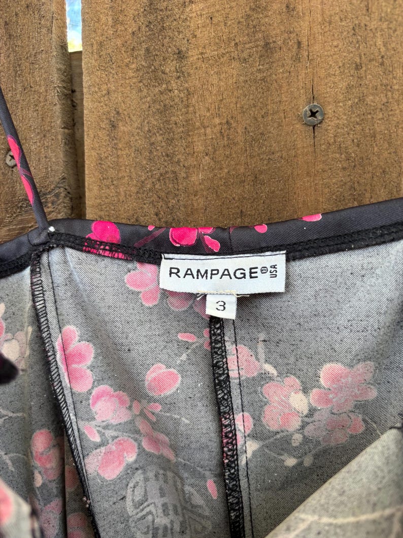 Op de afbeelding: Close-up van een zwarte jurk met een roze bloemenpatroon. De jurk heeft dunne bandjes en een V-hals. Het label leest "RAMPAGE USA" en maat "3". De stof heeft een subtiele textuur.