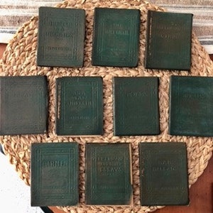 May include: A set of ten small, green leather-bound books with gold lettering on the spines. The books are arranged in two rows of five on a woven surface.