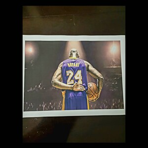 May include: A basketball player wearing a purple and gold jersey with the number 24 on the back, holding a basketball. The player is standing on a basketball court with a crowd in the background.