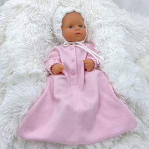 May include: A baby doll dressed in a pale pink sleep sack and a white knitted bonnet. The doll has blue eyes and rests on a fluffy white blanket. The sleep sack features three pink buttons down the front.