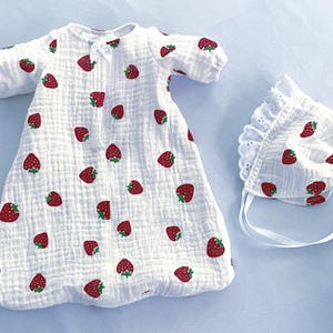 May include: A white long-sleeved gown featuring a repeating red strawberry print, a small white bow at the neckline, and elasticized sleeve cuffs. Accompanied by a matching white bonnet with a scalloped eyelet lace trim and white ribbon ties.