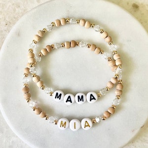 May include: Two stretch bracelets featuring light brown wooden beads, clear faceted crystal beads, and small gold-tone metallic spacers. One bracelet displays white round beads with black text reading MAMA, while the other features white beads with gold text reading MIA.