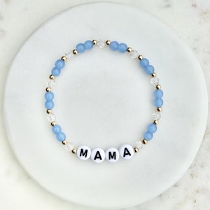 May include: A beaded bracelet with the word "MAMA" spelled out in white letter beads. The bracelet features light blue and clear beads, interspersed with gold-colored accents. The bracelet is displayed on a white, marble-like surface.