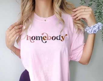 May include: A pink t-shirt with the word "homebody" printed in a stylized font with brown, pink, and gray colors.