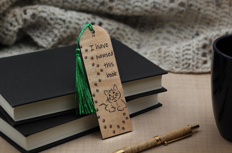 May include: Wooden bookmark with a green tassel, featuring a cat illustration and the text "I have pawsed this book".