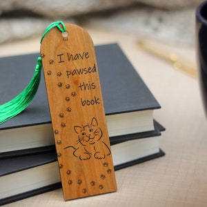 May include: Wooden bookmark with a green tassel. The bookmark has a cat illustration and the text "I have pawsed this book".