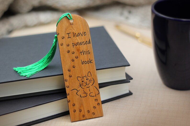 May include: Wooden bookmark with a green tassel. The bookmark has a cat illustration and the text "I have pawsed this book".