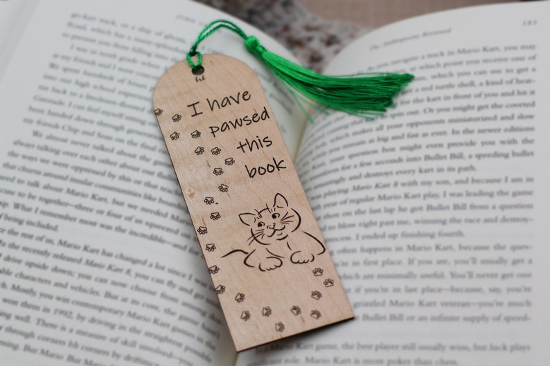 May include: A wooden bookmark with a green tassel. The bookmark features a cat illustration and the text "I have pawsed this book".