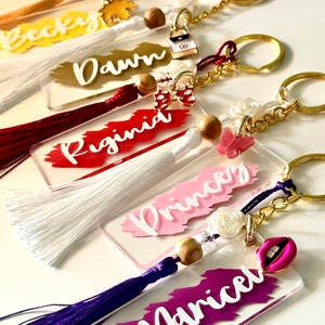May include: Acrylic keychains with names in white script on colorful brushstroke backgrounds. Each keychain has a tassel, gold-tone hardware, and decorative charms like a butterfly, lips, or a purse.