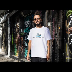 May include: White t-shirt featuring a small illustration of a boat and the phrase "The implication." The shirt is worn by a person standing in front of a graffiti-covered wall. The shirt is made of a soft, comfortable material.