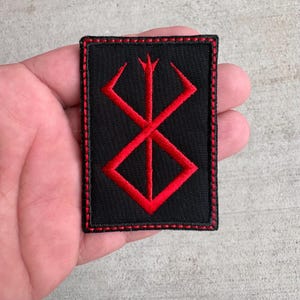 May include: A rectangular black embroidered patch featuring a bold red symbol of the Brand of Sacrifice. The patch has a thick red stitched border that frames the black fabric background, creating a high-contrast design with clean, dense embroidery.