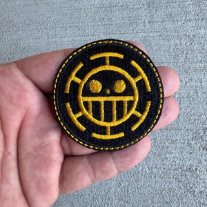 May include: A round, black embroidered patch with a yellow emblem. The emblem features a stylized face with a smile and a cross-like design around it. The patch is likely for clothing or accessories.