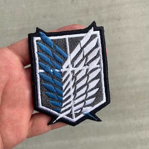 May include: A black and white embroidered patch with a blue and white wing design. The patch is shaped like a shield and has a black border.