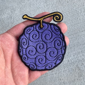 May include: A purple and black embroidered patch of a fruit with a golden stem. The fruit is round and has a swirl pattern.