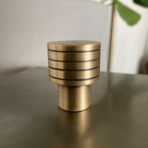 May include: A gold-toned metal knob with a cylindrical base and a top with four horizontal grooves.