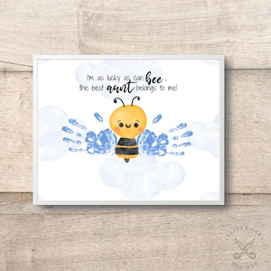 Puede incluir: Impresión enmarcada con una abeja de dibujos animados y el texto "I'm as lucky as can bee, the best aunt belongs to me!" La abeja es amarilla y negra, con alas de huellas de manos azules y acentos de nubes.