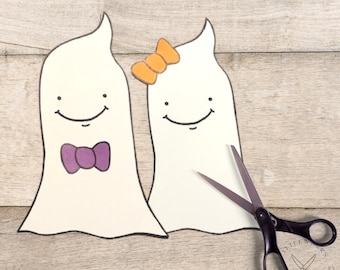 May include: Two white ghost cutouts with smiling faces, one with a purple bow tie and the other with an orange bow. A pair of scissors is next to the cutouts. The background is a light brown wooden surface.