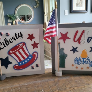 May include: Two wooden boxes decorated with patriotic themes. The box on the left has a red, white, and blue top hat with stars and the word "Liberty". The box on the right has a red, white, and blue star, the letters "USA", a Statue of Liberty silhouette, and the word "Liberty" with a torch.