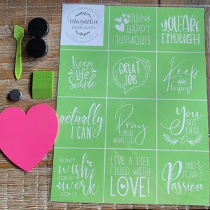 May include: A set of 10 green stencils with white lettering. The stencils feature positive affirmations such as "Think Happy Thoughts", "You Are Enough", "Keep Life Simple", "Great Job", "Keep Trying", "Actually I Can", "Pray More Worry Less", "You Got This", "Don't Wish For It Work For It", and "Live A Life Filled With Love!" The stencils are packaged with a pink heart-shaped sponge, two black paint pots, a green scraper, and a small black magnet.