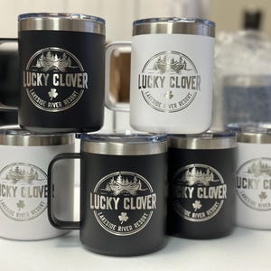 Puede incluir: Una colecci&oacute;n de seis tazas aisladas en blanco y negro con bordes plateados y tapas transparentes. Cada taza presenta un logotipo circular grabado con una canoa, &aacute;rboles y el texto Lucky Clover Lakeside River Resort con un tr&eacute;bol.