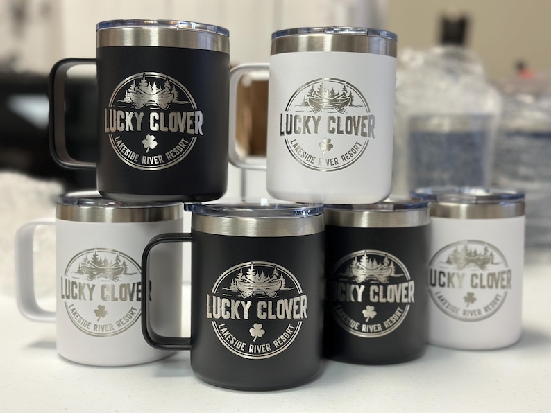 Puede incluir: Una colecci&oacute;n de seis tazas aisladas en blanco y negro con bordes plateados y tapas transparentes. Cada taza presenta un logotipo circular grabado con una canoa, &aacute;rboles y el texto Lucky Clover Lakeside River Resort con un tr&eacute;bol.