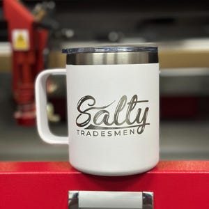 Puede incluir: Una taza t&eacute;rmica blanca con asa y tapa transparente. La taza presenta el texto Salty TRADESMEN grabado en una fuente oscura en la parte frontal. El cuerpo de la taza tiene un acabado texturizado y un borde de acero inoxidable.