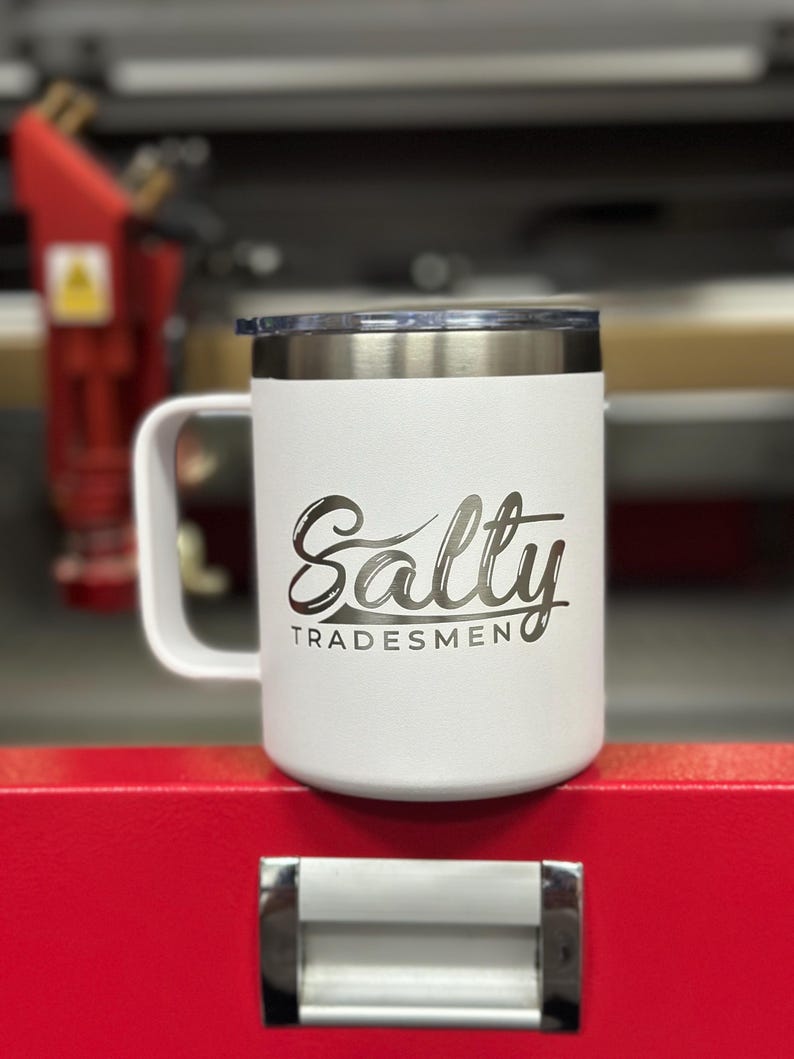 Puede incluir: Una taza t&eacute;rmica blanca con asa y tapa transparente. La taza presenta el texto Salty TRADESMEN grabado en una fuente oscura en la parte frontal. El cuerpo de la taza tiene un acabado texturizado y un borde de acero inoxidable.