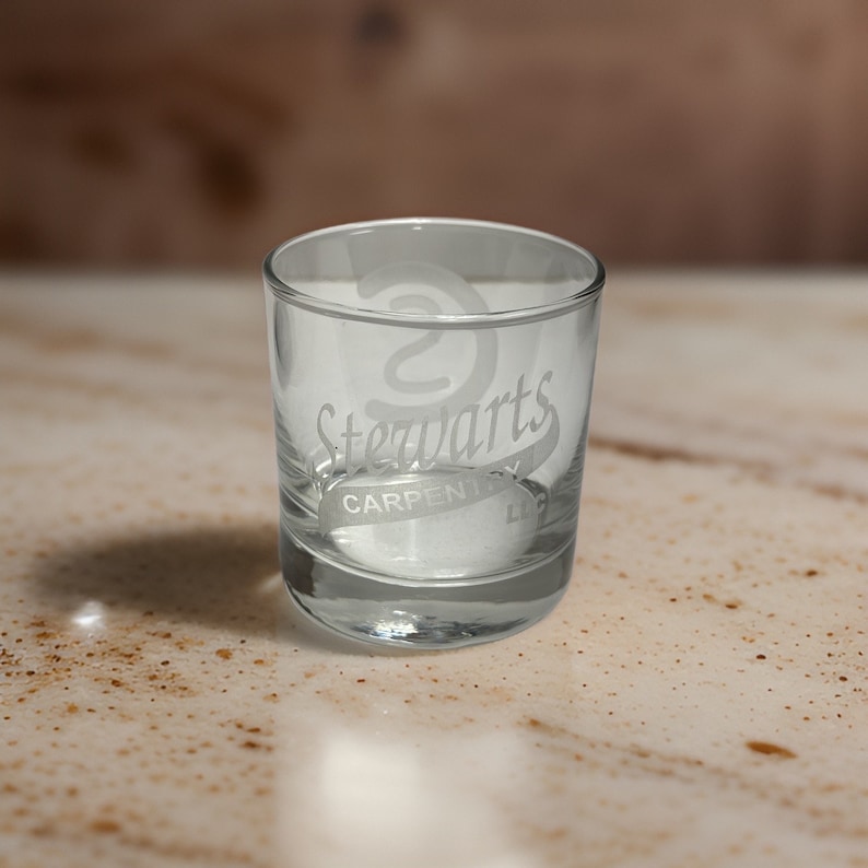 May include: A clear glass tumbler featuring an etched logo on the front. The design includes a stylized letter S above the text Stewarts, with the word CARPENTRY and the abbreviation LLC printed in a horizontal band underneath the main name.