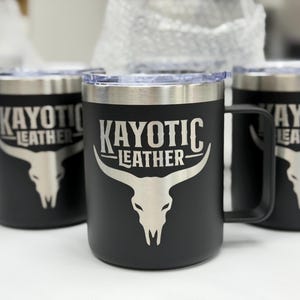 Puede incluir: Una taza de acero inoxidable negro mate con tapa transparente y asa. El frente presenta un gr&aacute;fico grabado con l&aacute;ser en tono plateado de un cr&aacute;neo de buey longhorn bajo el texto KAYOTIC LEATHER en una fuente serif estilizada.