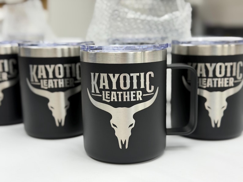 Puede incluir: Una taza de acero inoxidable negro mate con tapa transparente y asa. El frente presenta un gr&aacute;fico grabado con l&aacute;ser en tono plateado de un cr&aacute;neo de buey longhorn bajo el texto KAYOTIC LEATHER en una fuente serif estilizada.